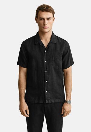 Jonge man met een zwart overhemd met korte mouwen en bijpassende zwarte broek, staand tegen een effen witte achtergrond.