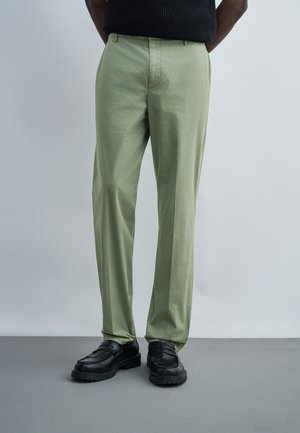 Personne portant un pantalon vert clair à jambes droites et des mocassins en cuir noir, debout devant un fond uni gris clair.