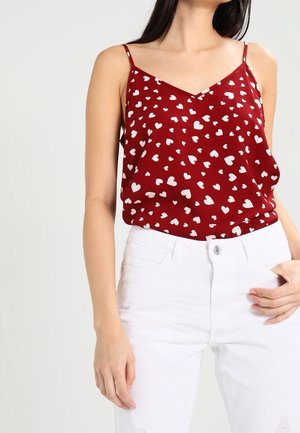 Vrouw draagt een rood mouwloos topje met witte hartpatronen en witte broek, staat met één hand in de zak tegen een effen achtergrond.