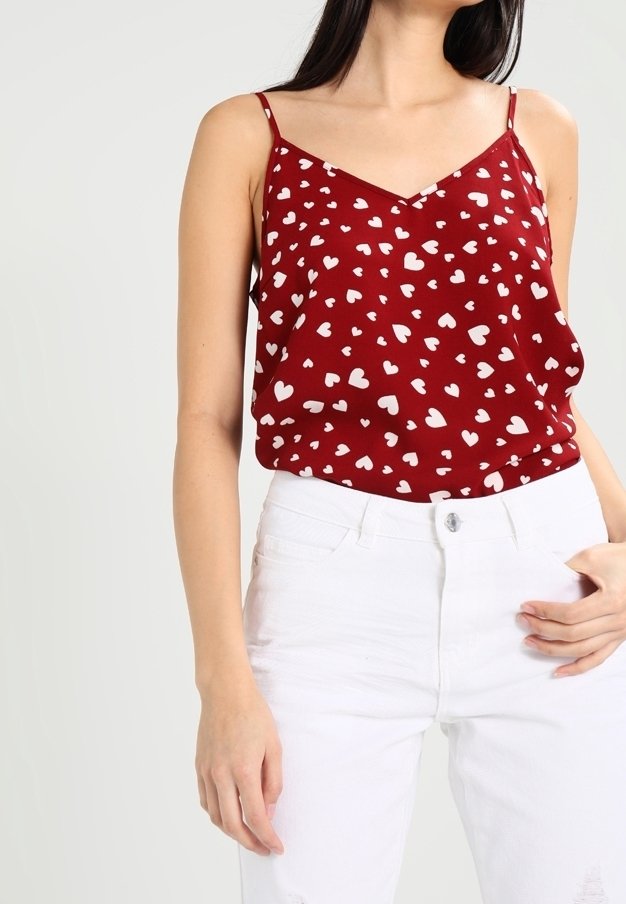 Femme portant un haut sans manches rouge avec des motifs de cœurs blancs et un pantalon blanc, debout, une main dans la poche, sur un fond uni.