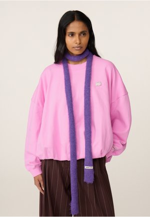 Person trägt einen knallpinkfarbenen übergroßen Pullover, einen langen violetten Schal, der einmal um den Hals gewickelt ist, und braune Nadelstreifenhose.