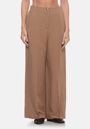 Pantaloni marroni a gamba larga, stretti davanti con bottone e zip, indossati con scarpe marrone scuro su sfondo chiaro.