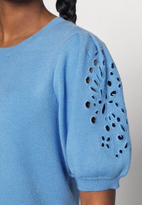 Maglione a maglia azzurro chiaro con maniche a sbuffo corte, caratterizzato da fantasie floreali decorative traforate sulle maniche e un collo a costine.