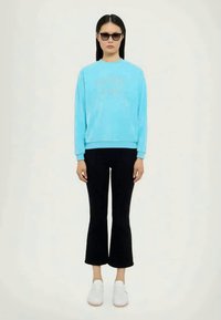 Tyrkysová mikina s kulatým výstřihem a dlouhými rukávy, spárovaná s černými rozšířenými kalhotami a bílými teniskami. Celkový vzhled doplňují sluneční brýle.