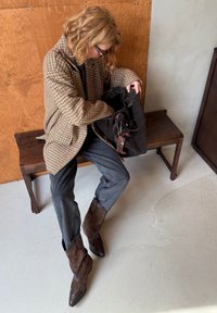 Brązowa marynarka w kratę, czarna zamszowa torebka typu bucket, szare dżinsy i brązowe zamszowe spiczaste botki z gwiazdkowymi zdobieniami, siedzące na ciemnym drewnianym ławce.