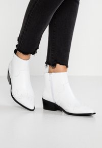 Botas de tobillo de cuero blanco con cremalleras laterales, combinadas con jeans negros con dobladillo deshilachado, posando sobre un suelo blanco liso.