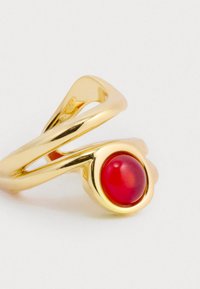 Zlatý prsteň s prekrúteným dizajnom, ktorý má stredový okrúhly červený kameň. Hladký povrch, lesklá textura a moderný, minimalistický tvar.