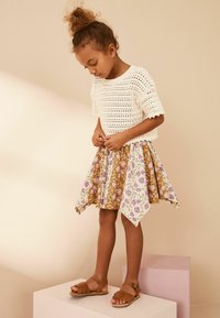 Top bianco a maglia con maniche corte, abbinato a una gonna floreale nei toni del beige e del lilla. Sandali con cinturini color nocciola completano l'outfit.