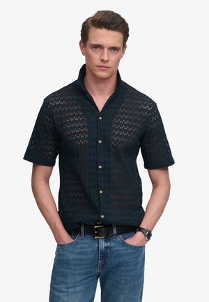Junger Mann trägt ein kurzärmeliges, schwarzes, halbtransparent gemustertes Hemd mit blauen Jeans und schwarzem Gürtel, Hände in den Taschen.