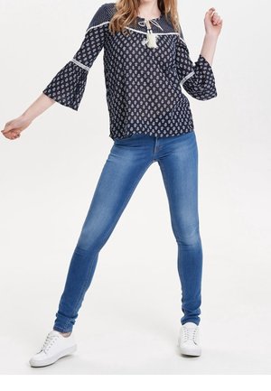 Frau trägt eine marineblaue gemusterte Bluse mit Glockenärmel, blaue Skinny-Jeans und weiße Sneakers, posiert mit auseinanderstehenden Beinen und erhobenen Armen.