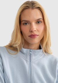 Gaiši zila rāvējslēdzēja jaka ar augstu apkakli, izgatavota no mīksta auduma ar redzamām šuvēm. Rāvējslēdzēja aizdare ar kvadrātveida vilkšanas cilpu.