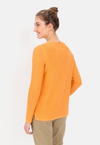 Pomarańczowy sweter z długim rękawem o prążkowanej teksturze, okrągłym dekolcie i bocznymi rozcięciami u dołu, noszony na beżowych spodniach.