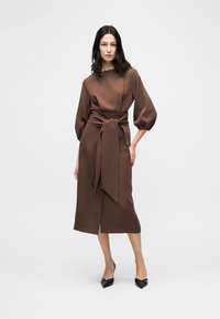 Vrouw in een bruine midi-jurk met pofmouwen en een brede, gestrikte tailleband, draagend zwarte pumps met een puntige neus, staand tegen een effen achtergrond.