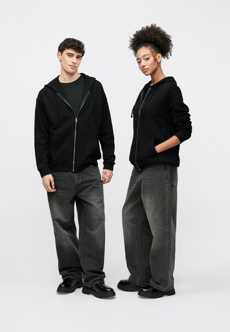 Felpe con cappuccio nere con zip e tasche frontali, indossate sopra maglie scure. Entrambi indossano jeans grigi a gamba larga e scarpe nere robuste. Outfit semplice e casual.