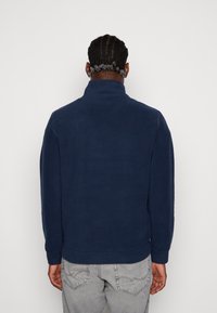 Giacca in pile blu navy con colletto alto, texture morbida e vestibilità comoda, abbinata a jeans grigio chiaro con dettagli di cucitura sulle tasche posteriori.