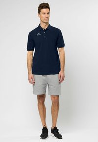 Marineblauw poloshirt met een drieknops knoopsluiting en logo, gecombineerd met grijze shorts van zacht materiaal. Het model draagt zwarte sportschoenen.