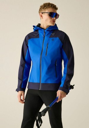 Man met een blauw-zwarte outdoorjas en zonnebril die een blauwe wandelstok vasthoudt tegen een effen achtergrond.