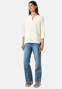 Witte blouse met driekwart mouwen, een diepe halslijn en knoopdetails, gecombineerd met lichtblauwe jeans en platte sandalen.