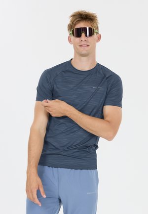 Homme portant un t-shirt de sport foncé à motifs, un short bleu clair et des lunettes de sport réfléchissantes, ajustant sa manche gauche sur fond blanc.