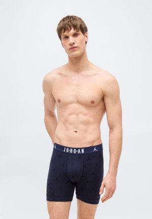 Jeune homme aux cheveux bruns courts portant un boxer Jordan bleu marine avec ceinture à logo, debout devant un fond clair uni.