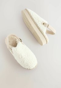 Des chaussures blanches et duveteuses à enfiler, avec un dessus texturé et une bande élastique beige. La semelle est légère et présente un motif de bande de roulement en zigzag.