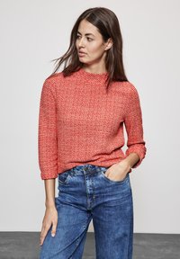Maglione rosso a trame lavorate con collo alto e maniche a tre quarti, abbinato a jeans in denim a vita alta di colore azzurro chiaro.