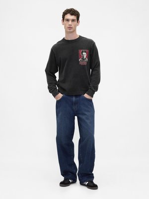 Μαύρο μακρυμάνικο πουκάμισο με γραφικό εκτύπωμα στο στήθος, συνδυασμένο με φαρδιά μπλε τζιν και μαύρα αθλητικά παπούτσια σε απλό φόντο.