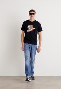 Tricou grafic negru cu un design de leu și textul "Barbour", asortat cu blugi albaștri deschiși și teniși negri.