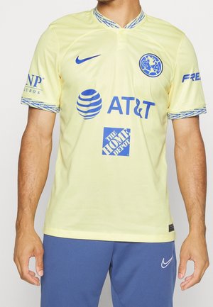 Homme portant un maillot de soccer jaune du Club América avec des logos bleus incluant Nike, AT&T, The Home Depot, et un short bleu avec logo Nike blanc.
