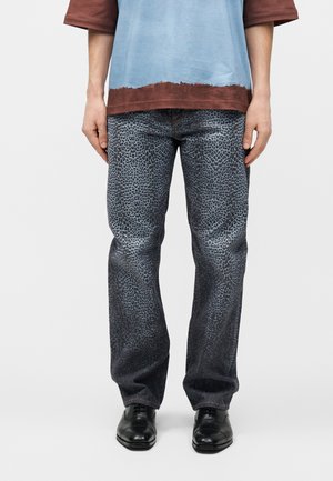 Άτομο που φοράει φαρδιά γκρι παντελόνια με λεοπάρ σχέδιο, μαύρα επίσημα παπούτσια και μπλε-καφέ μακρυμάνικο πουκάμισο, στέκεται σε λευκό φόντο.