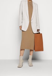 Blazer de doble chaleco en un color beige claro sobre un vestido ajustado marrón. Botines grises hasta el tobillo, llevando un bolso tote rectangular marrón. Texturas suaves.
