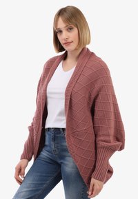 Junge Frau, die eine mauvefarbene Strickjacke mit Rautenmuster über einem weißen T-Shirt und blauen Jeans trägt, steht vor einem weißen Hintergrund.