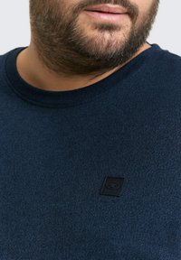 Dunkelblaues, strukturiertes T-Shirt mit Rundhalsausschnitt, das ein kleines schwarzes Logopatch auf der Brust aufweist. Weicher Stoff mit lässiger Passform.