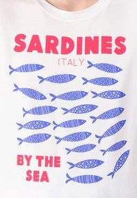 T-shirt branca com um padrão de peixes azuis com diversos designs, e texto vermelho a dizer "SARDINHAS ITÁLIA À BEIRA-MAR."