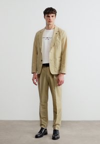 Νέος άνδρας με μπεζ κοστούμι, λευκό μπλουζάκι με σχέδιο και μαύρα παπούτσια, στέκεται μπροστά σε απλό λευκό τοίχο.