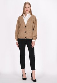 Cardigan bej tricotat cu decolteu în V, croi oversized, și nasturi negri, asortat cu pantaloni negri mulați și pantofi cu toc înalt negri.