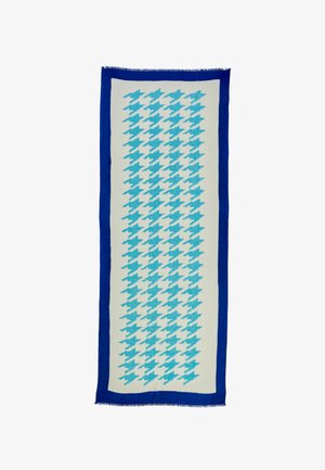 Let tørklæde med blåt og hvidt geometrisk mønster, frynset kant, draperet over en grå top. Kombineret med mørke bukser med vide ben.