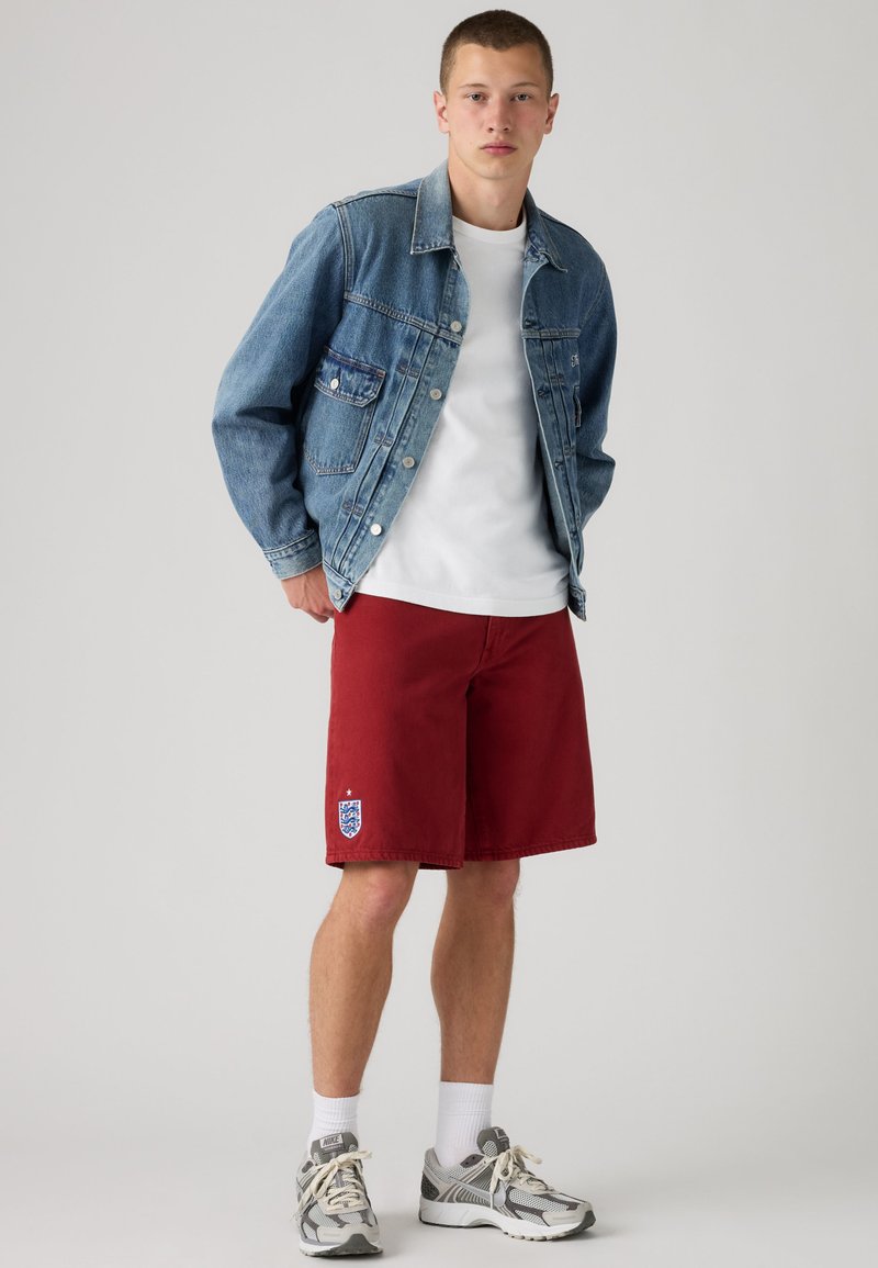 Joven con chaqueta vaquera azul, camiseta blanca, pantalones cortos rojos con el emblema de Inglaterra, calcetines blancos y zapatillas Nike grises, de pie frente a un fondo liso.