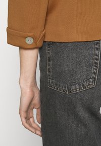 Rękaw z brązowej dzianiny z podwiniętym mankietem i okrągłym guzikiem. Szare jeansy z tylną kieszenią. Faktura wydaje się szorstka.