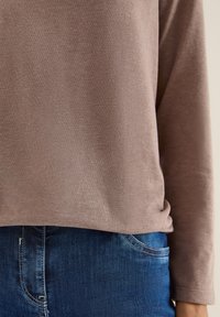 Camicia a maniche lunghe in tessuto marrone chiaro con una texture liscia, abbinata a jeans in denim blu scuro con accenti di cucitura.