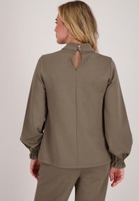 Vrouw in een taupe lange mouw top met een hoge kraag en een keyhole opening aan de achterkant, vastgemaakt met twee knoppen, gecombineerd met bijpassende broeken.