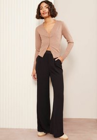 Lichtbruin cardigan met een v-hals en gouden knopen, gecombineerd met zwarte wijde broek en beige schoenen, tegen een neutrale achtergrond.