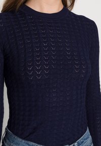 Navy knit long-sleeve top featuring a textured diamond pattern, crew neckline, and a fitted silhouette. Worn with a light denim waistband.
