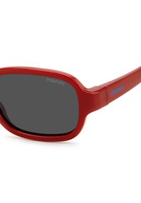 Lunettes de soleil en plastique rouge au design épuré, avec des verres gris foncé et un détail de logo subtil sur la branche latérale. Finition lisse et brillante.