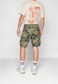 Mies, joka pukeutunut beigeen graafiseen t-paitaan, vihreisiin maastokuvioisiin cargo shortseihin, valkoisiin Adidas-tennareihin ja valkoisiin sukkiin, joissa on sinisiä ja ruskeita raitoja, seisoo sis ällä.