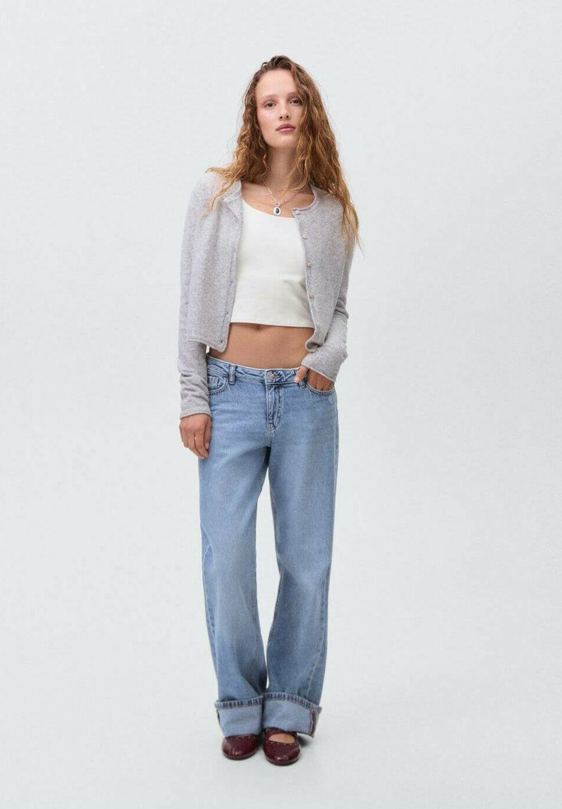 Světle šedý pletený kardigan přes bílý crop top, v kombinaci s volně střiženými modrými džíny a hnědými botami. Jednoduchý design s ležérním stylem.