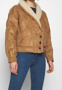 Brun mocka jacka med en fluffig krämfärgad sjalkraga, fyra knappar och sidofickor. Klippt i en kort, fyrkantig design.