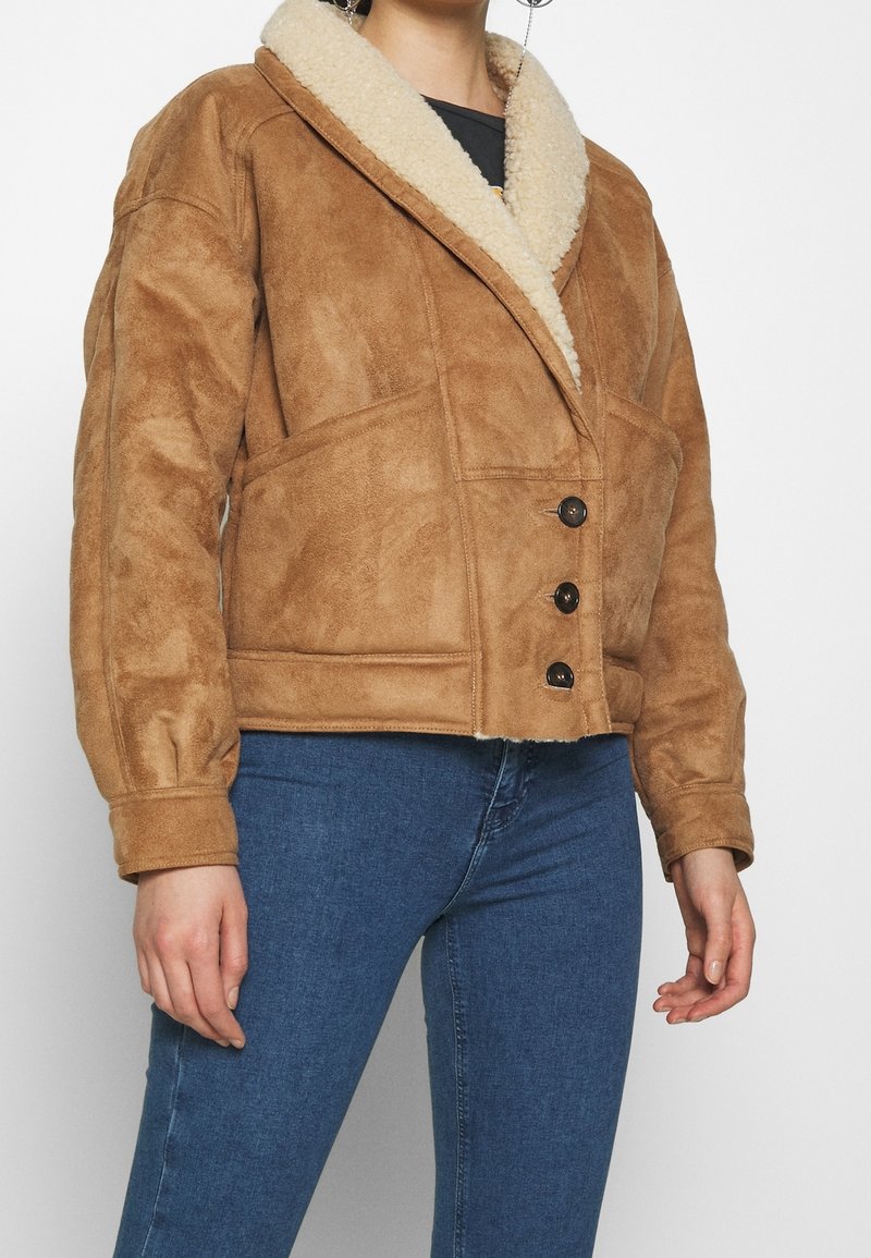 Brun mocka jacka med en fluffig krämfärgad sjalkraga, fyra knappar och sidofickor. Klippt i en kort, fyrkantig design.