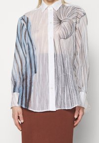 Genomskinlig blus med lång ärm som har abstrakta mönster i blått och svart, plisserad textur och en vit knapp-up framtill.