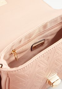 Kabelka v broskvové barvě s texturovaným geometrickým vzorem, obsahující zipovou přihrádku, zlaté kovové detaily a hladké vnitřní podšívku.